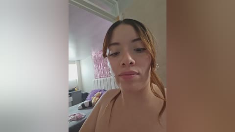 A video of a young, light-skinned woman with straight, light brown hair in a ponytail, wearing a beige top, standing in a bedroom with pink and purple decorations.