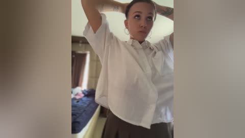 A video of a woman with short brown hair, wearing a white blouse and dark skirt, adjusting her hair in a bedroom with a bed and messy blue sheets in the background.