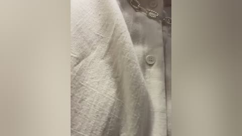 A close-up video of a person's chest, partially covered by a white, textured shirt with a single button, showcasing a silver chain necklace. The background is blurred, focusing on the textures and details of the shirt.