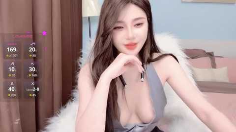 Video of an East Asian woman with long, straight brown hair, fair skin, and pink lipstick. She's wearing a low-cut gray top, exposing cleavage, and sits on a white fur blanket. Background shows a pastel-colored bedroom with a bed and curtains.