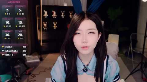 Video of a young Asian woman with long black hair and blue bunny ears, wearing a light blue top, in a dimly lit room with a computer screen displaying live streaming statistics.