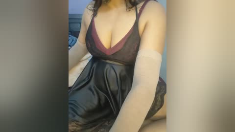 A video of a curvy, light-skinned woman with dark hair, wearing a sheer, black lingerie dress with a deep V-neck and sheer white stockings. She sits on a bed in a dimly lit room.