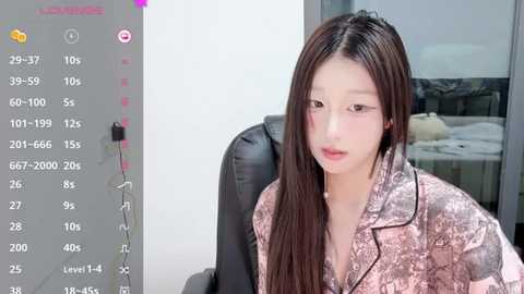 Video of an East Asian woman with long black hair, wearing a pink floral blouse, seated in a black leather chair. Background shows a white wall and a bed.