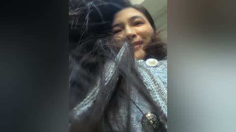 A video of a woman with long black hair, wearing a gray fur coat with a silver button, and a silver necklace with a pendant. Her face is partially obscured by her hair, which is blowing in the wind.
