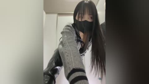 Video of an Asian woman with long black hair, wearing a black face mask, gray jacket, and knee-high boots, walking down a dimly lit hallway.