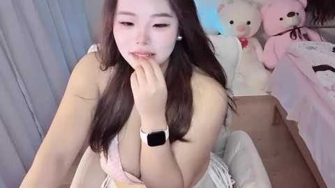 Video of an East Asian woman with long black hair, wearing a pink bra, sitting on a bed. She's touching her lips with a finger. The room has soft pastel tones and stuffed animals.