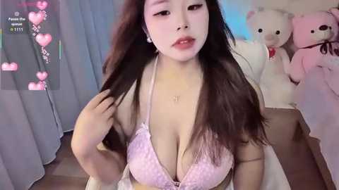 A video of an East Asian woman with long, straight black hair, wearing a pink lace bra, sitting on a bed with plush toys.