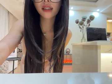 Video of an Asian woman with long, straight black hair and glasses, wearing a floral top, sitting in a modern living room with beige walls, a TV, and decorative flowers.