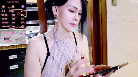 Video of a slender, light-skinned Asian woman with long black hair in a bun, wearing a delicate lace top, seated indoors, focused on a tablet.