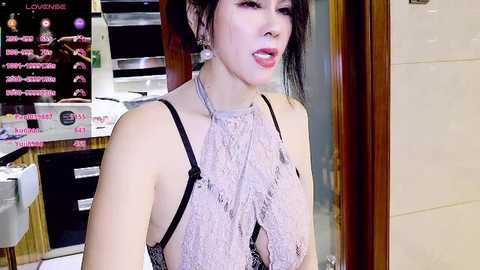 Video of an East Asian woman with pale skin and dark hair, wearing a sheer, lace top and dangling earrings, standing indoors next to a glass door, surrounded by restaurant signs.