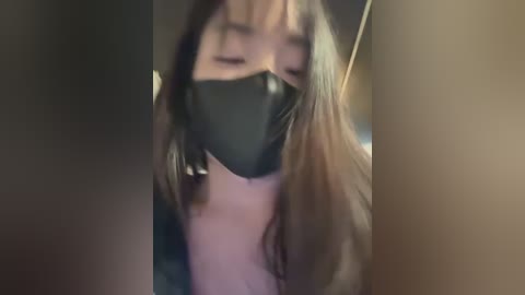 Video of a young Asian woman with long, straight brown hair and fair skin, wearing a black face mask, partially obscured by two large, dark objects.
