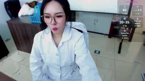 Video of an Asian woman with long black hair, wearing glasses and a white lab coat, sitting in a cluttered office with a desk, shelves, and a blue basket in the background.