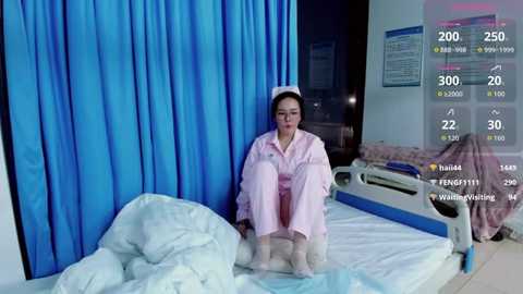 A video of an Asian woman in a pink hospital gown sitting on a bed, surrounded by blue curtains, medical equipment, and a digital screen displaying vital signs.