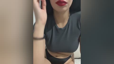 A video of a light-skinned woman with long black hair, wearing a tight grey crop top and high-waisted black panties, leaning against a beige wall, with a subtle tattoo on her left arm.