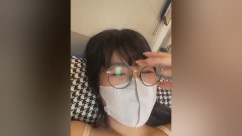 A video of a woman with long black hair, wearing a white mask and round glasses, being touched by another hand on her face. She lies on a black-and-white houndstooth-patterned blanket, in a dimly lit room.