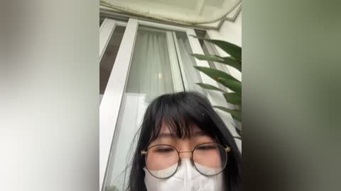 Video of an Asian woman with long black hair, wearing round glasses and a white mask, standing indoors near a glass door with vertical blinds.