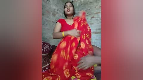 Video of a South Asian woman in a vibrant red sari with golden patterns, sitting on a bed, against a rustic stone wall background.
