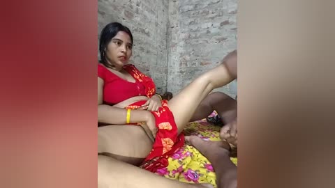 Video of a South Asian woman with medium skin tone, wearing a red and yellow floral sari, reclining on a colorful bed. Background shows a rustic, brick-walled room.
