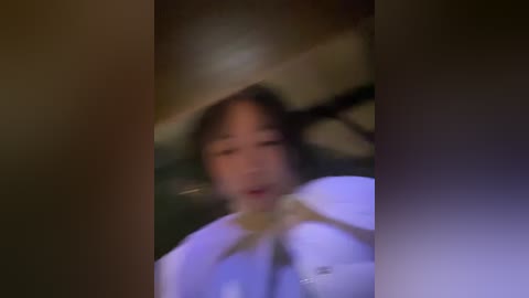 Blurry video of an Asian woman with shoulder-length black hair, wearing a white dress, and a large white hat. The background is dimly lit with indistinct objects.