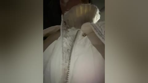 A close-up video of a woman's torso, focused on her chest, with her right hand adjusting a white, zipped, fluffy jacket. The background is dimly lit, creating a shadowy effect.
