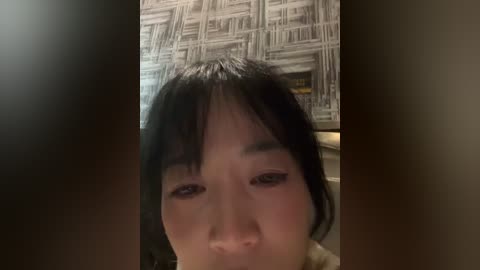 A close-up video of a young East Asian woman with dark hair and bangs, wearing dark eye makeup, taken indoors with a blurred background featuring a grey and white abstract patterned wall.