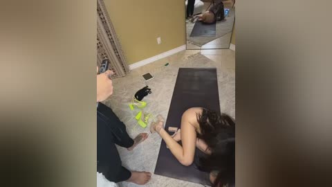 A candid video shows a woman in a dark room, kneeling on a yoga mat, facing a mirror. She wears a black bra, holding a phone and looking at her reflection. A man stands beside her, wearing a dark shirt.