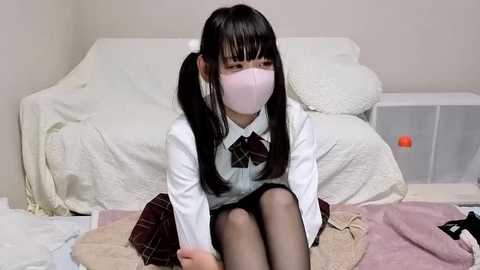 Video of an Asian girl with long black pigtails, wearing a white mask, school uniform, and a plaid skirt, sitting on a bed with a white blanket, and a red box in the background.