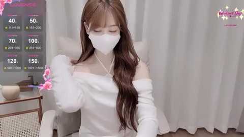 A video of an Asian woman with long brown hair, wearing a white off-shoulder top and face mask, adjusting her hair in a home setting with a white curtain backdrop.