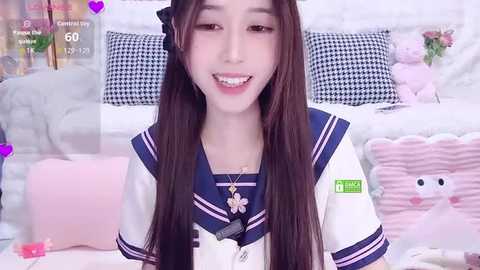 Video of an Asian woman with long brown hair in a schoolgirl outfit, smiling, surrounded by pastel pink and white bedding with decorative pillows.