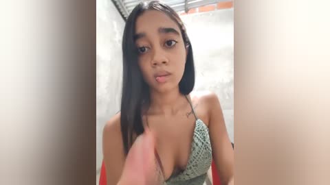 A video of a young, dark-skinned girl with long, straight black hair, wearing a low-cut, green patterned top. She looks serious, standing in a dimly lit room with a brick wall and red curtains in the background.