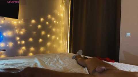 Media: Video of a nude man lying on a bed, wearing a white hat, with a soft glow of string lights in the background.