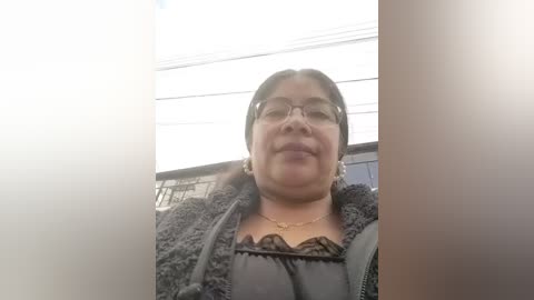 Video of a middle-aged woman with medium-dark skin and glasses, wearing a black lace top and a dark fur jacket. She has a neutral expression and is standing in front of a blurred background with windows.