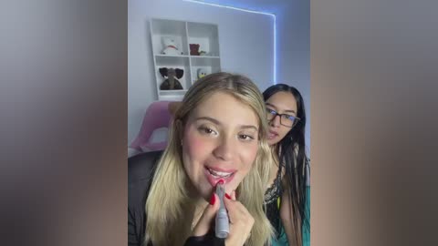 Video of two women, one with blonde hair and the other with black hair and glasses, applying lipstick in a modern bedroom with white shelves and blue LED lights.