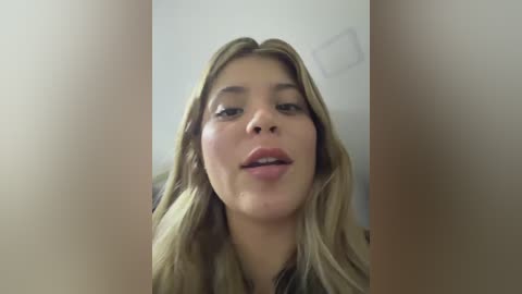 A video of a young woman with fair skin, light blonde hair, and a neutral expression, captured from a slightly low angle, possibly indoors with a plain white wall as the background.