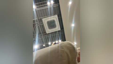 Video of a ceiling with a central HVAC unit, surrounded by string lights, taken from a low angle, showing a person's arm and beige sofa in the foreground.