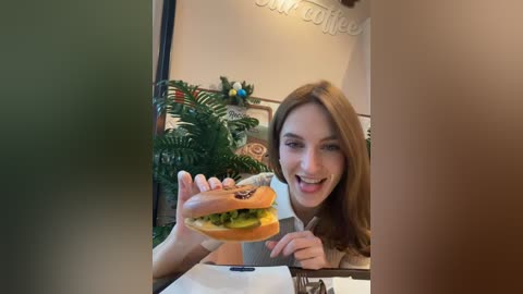 Video of a smiling, light-skinned woman with long brown hair, holding a hamburger in a cozy caf\u00e9. Background features greenery, decorative elements, and a \"Coffee\" sign.