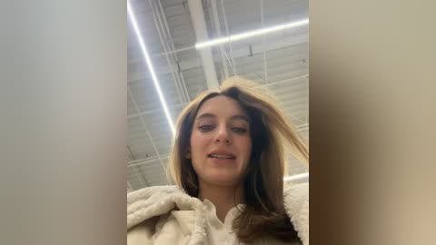 Video of a young woman with long brown hair, fair skin, and a casual beige jacket, captured from a low angle, in a dimly lit, industrial warehouse with exposed white beams and fluorescent lighting.