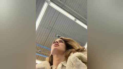 Video of a young Caucasian woman with fair skin and long blonde hair, wearing a beige coat, looking up with a contemplative expression. The background features a corrugated metal ceiling with fluorescent lights.
