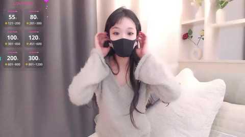 Video of an Asian woman with long black hair, wearing a black face mask, white fuzzy robe, and white headphones, in a cozy, bright living room.
