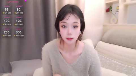 Video of an East Asian woman with fair skin, black hair, and small breasts, wearing a gray fuzzy sweater, seated on a beige couch in a softly lit room with a white shelf and flowers in the background.