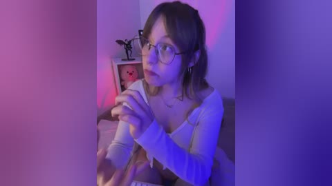 Video of a young woman with glasses, light skin, and brown hair, wearing a white long-sleeve top, sitting on a bed, holding a piece of toast, in a dimly lit room with purple lighting.