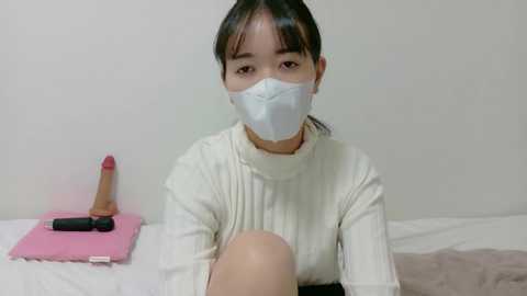 Video of an Asian woman with long black hair, wearing a white mask, a white sweater, and a pink blanket, sitting on a bed.