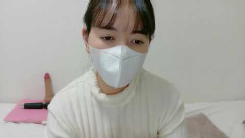 Video of a young East Asian woman with dark hair, wearing a white face mask, light sweater, and seated on a white bed with a pink pillow.