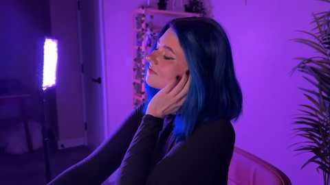 Video of a fair-skinned woman with vibrant blue hair, wearing a black top, seated with her hands on her face, illuminated by a purple light, in a dimly lit room.