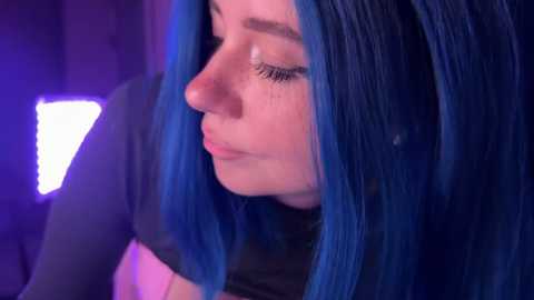 Video of a fair-skinned woman with blue hair, wearing a black top, looking downwards in a dimly lit room with purple and blue lighting.