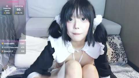 A young Asian woman with straight black hair in pigtails, dressed as a French maid, lies on a bed with patterned pillows. The room has gray walls and a white couch.