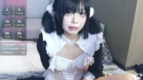 Video of a young Asian woman with black pigtails in a revealing white and black maid costume, sitting on a bed with a gray headboard, displaying live stream stats in the corner.