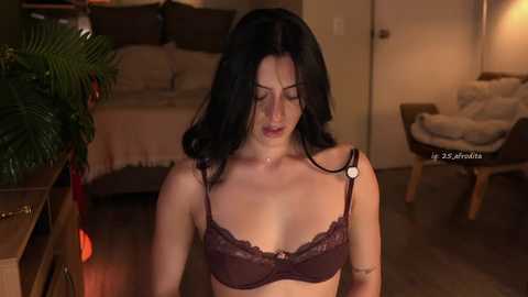 Video of a fair-skinned woman with long black hair, wearing a maroon lace bra, in a dimly-lit living room with a bed, chair, and plant.