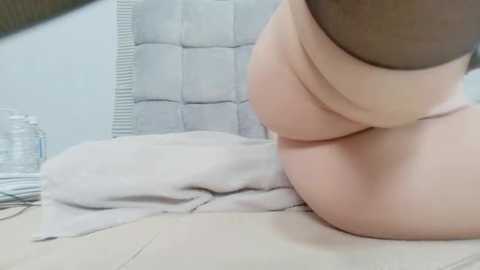 Video of a person with light skin, sitting on a cream-colored bed, wearing black thigh-high stockings and beige panties. The background features a white, tufted headboard and a glass jar on a bedside table.