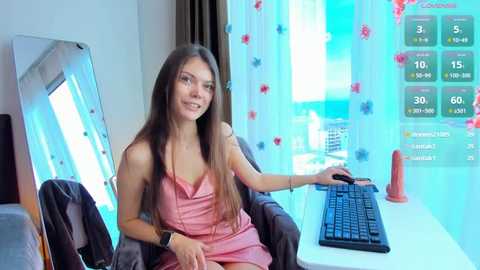 Video of a smiling, slender woman with long brown hair in a pink dress, sitting at a desk with a computer, a keyboard, and an orange dildo.